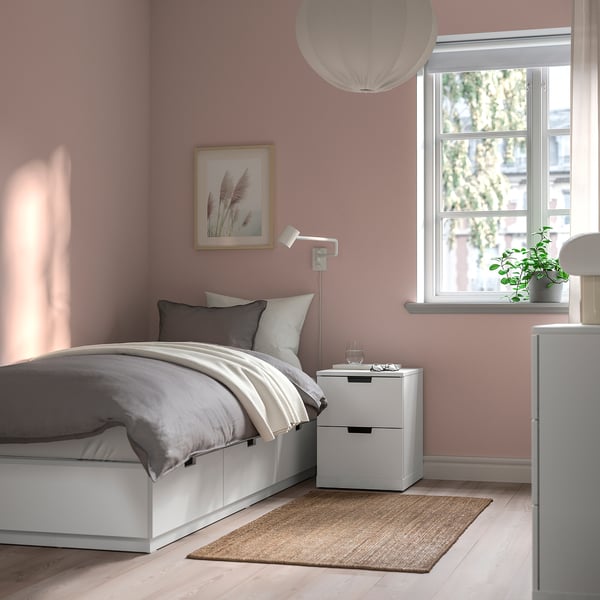 Camera da letto minimalista con letto NORDLI bianco, biancheria da letto grigia, comodino, lampada, tappeto. Pareti rosa, grande finestra per una luminosità accogliente.