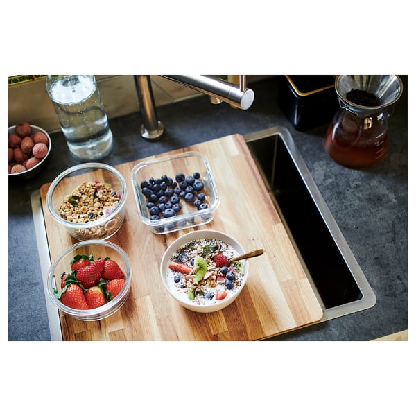 Tagliere da cucina in legno con ciotole di frutta e muesli su un piano di lavoro scuro.