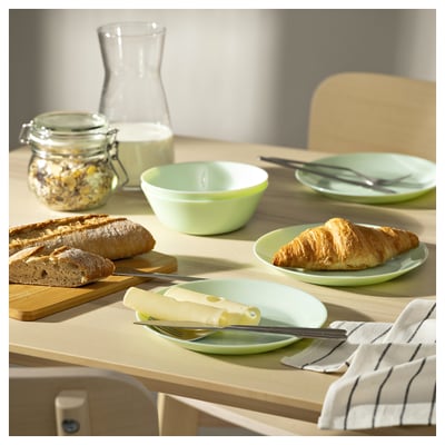 Set di stoviglie in vetro verde brillante su tavolo in legno, con ciotola, piatto con pane e coltello da formaggio. Allestimento colazione fresco e invitante.