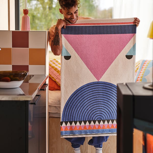 Persona che tiene in mano un grande, colorato, geometrico cotone OMMJÄNGE appeso a parete, più grande degli oggetti da cucina vicini.