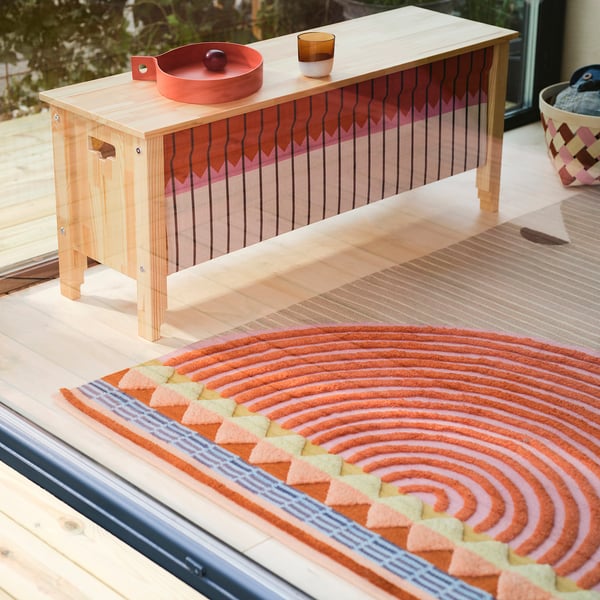 Grande tappeto rustico OMMJÄNGE con disegno geometrico in lana sotto una panca in legno chiaro vicino alla finestra, che mostra motivi e trame intricati.