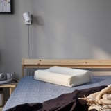 Una camera da letto minimalista, un cuscino PAPEGOJBUSKE su un letto con testiera in legno, una lampada moderna e un piccolo comodino con cuffie.