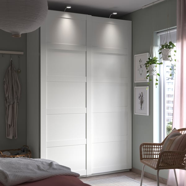 Camera da letto moderna con armadio PAX bianco, piante sospese, sedia in vimini, letto con coperta a righe, pareti grigio chiaro e finestra con vista sulla città.