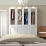 Camera da letto moderna con grande armadio PAX bianco, letto beige, sedia in vimini e luci soffuse.