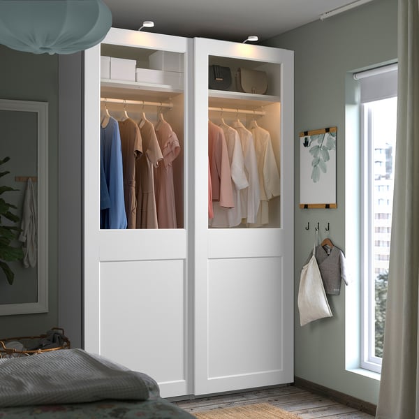 Camera da letto moderna dotata di un grande armadio PAX bianco con ante in vetro che mostrano i vestiti, situato accanto a una grande finestra con tende.