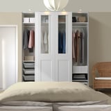 Un grande armadio PAX bianco con vetro, che espone vestiti e oggetti organizzati. Si erge alto accanto a una sedia di vimini e un letto con biancheria da letto beige.