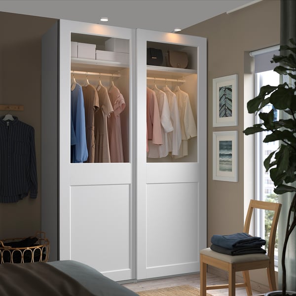 L'armadio moderno con ante in vetro mostra i vestiti, fondendosi con pareti beige e grandi finestre. Una sedia tiene accanto i vestiti piegati.