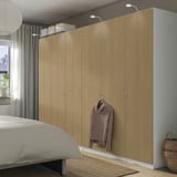 Camera da letto moderna con armadio PAX in legno chiaro, spazio per appendere, mensole, maglione marrone, biancheria da letto grigia, grande finestra e cesto intrecciato.