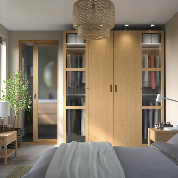 Camera da letto moderna con grande armadio in legno PAX; mensole e cassetti, affiancati da comodini con lampade. Illuminazione soffusa dal ciondolo intrecciato sopra.