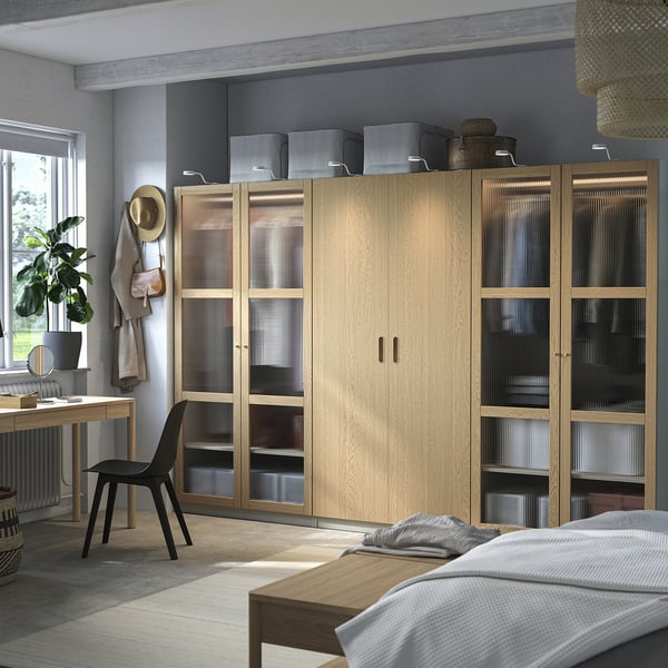 L'armadio PAX in rovere, dal design moderno, ampio e alto, ospita un contenitore con ante in vetro, si affianca a una scrivania e una sedia nera.