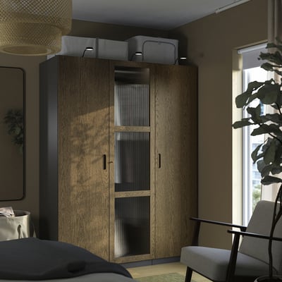 Una camera da letto moderna con un alto armadio in legno PAX, porte con chiusura ammortizzata e scatole portaoggetti grigie. Una finestra e una pianta aggiungono luce naturale e verde.