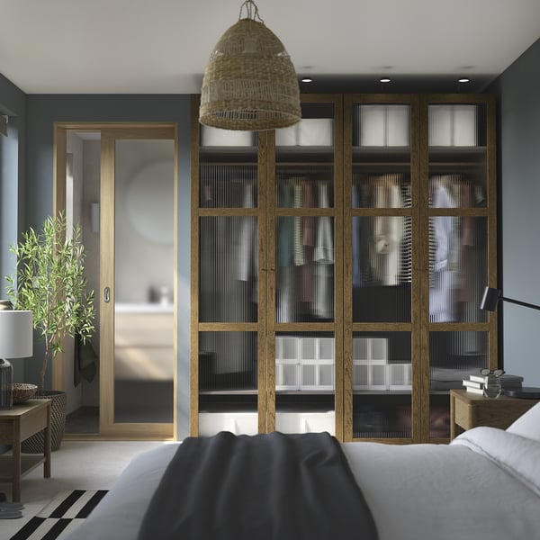 Camera da letto moderna con armadio alto in legno scuro PAX, pannelli in vetro, porta scorrevole in legno chiaro, lampada a sospensione e letto a righe.