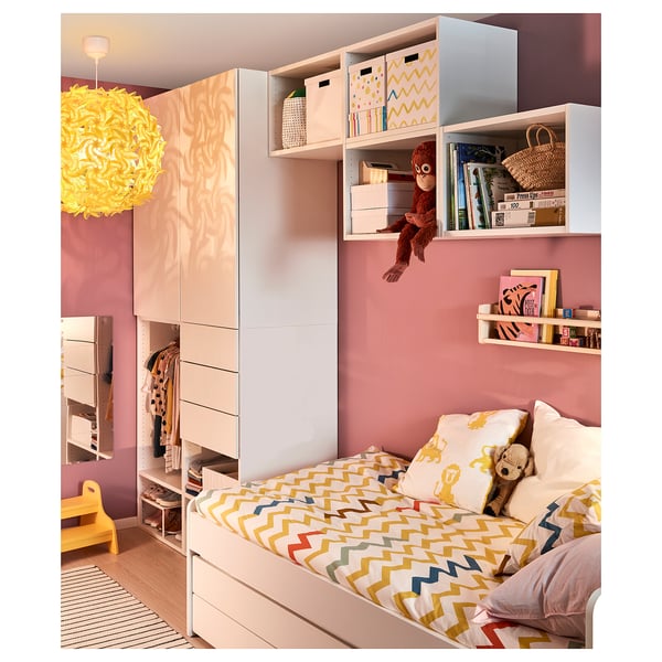 Camera da letto per bambini con pareti rosa, biancheria da letto vivace e un giocattolo a forma di scimmia appesa. Contenitori bianchi con scatole colorate e una lampada gialla.