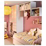Camera da letto per bambini con pareti rosa, biancheria da letto vivace e un giocattolo a forma di scimmia appesa. Contenitori bianchi con scatole colorate e una lampada gialla.