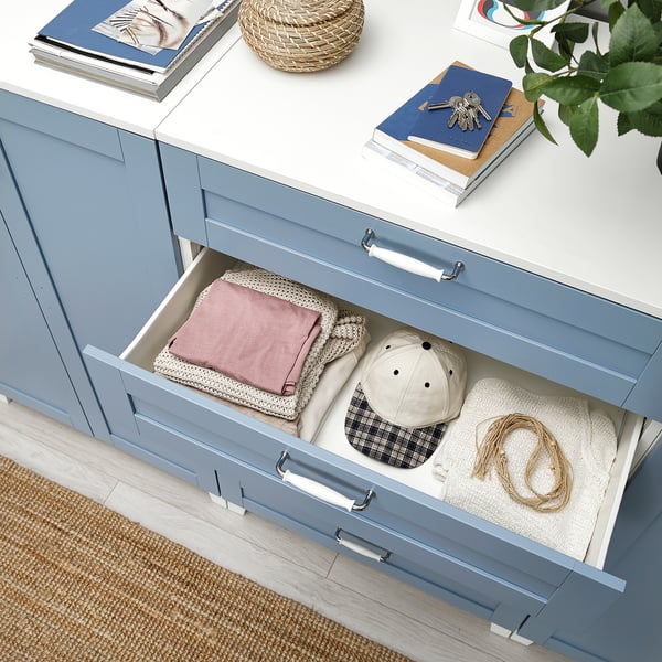 Cassettiera PLATSA blu con cassetto aperto che rivela abiti e accessori piegati. Top ha libri, chiavi e piante.