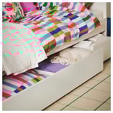 Biancheria da letto a righe luminose su letto bianco, con cassetto aperto che mostra i vestiti piegati, su tappeto beige e verde.