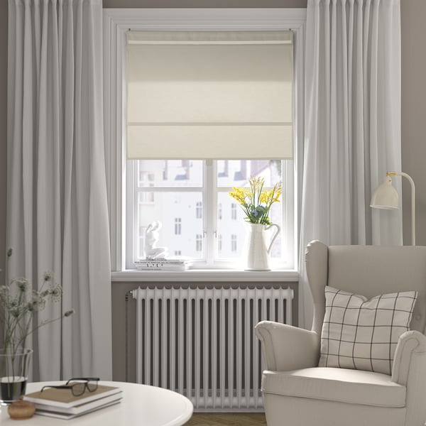 Angolo accogliente con tenda beige RINGBLOMMA, poltrona bianca e vaso di fiori gialli accanto a una finestra.
