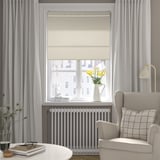Angolo accogliente con tenda beige RINGBLOMMA, poltrona bianca e vaso di fiori gialli accanto a una finestra.