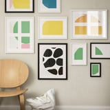 Cornici astratte colorate sulla parete con una centrale nera. Sedia in legno con cuscino bianco davanti.