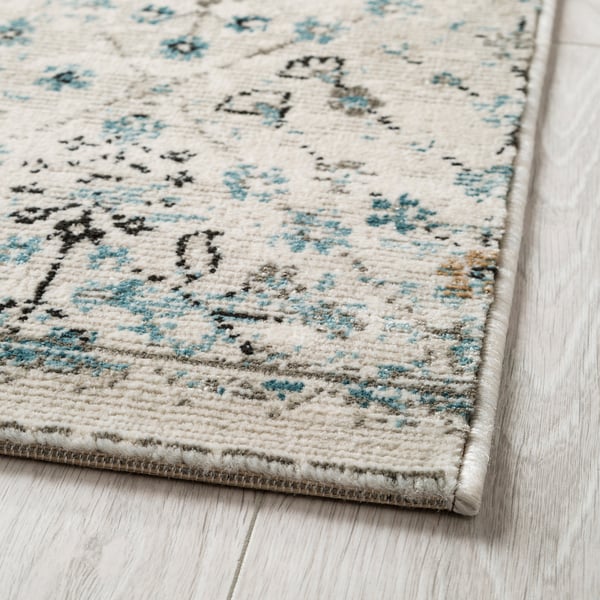 Tappeto di colore chiaro con motivi floreali, accenti bluastri e un pelo di peluche su un pavimento di legno.