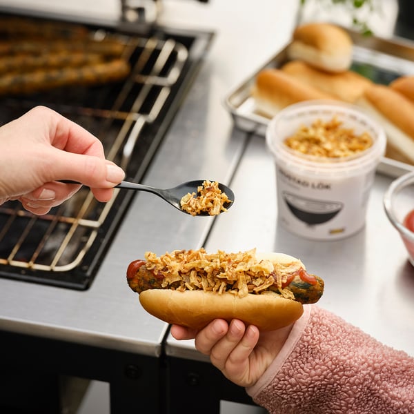 Una persona prepara un hot dog, aggiungendo condimenti di cipolla da un contenitore etichettato ROSTAD LÖK. L'impostazione della cucina mostra un fornello a gas e altri oggetti da cucina.