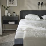 Scena della camera da letto con piumino SANDGRÄSMAL sul letto, comodino grigio che contiene libri, orologio e pianta; testiera nera, lampade da parete.