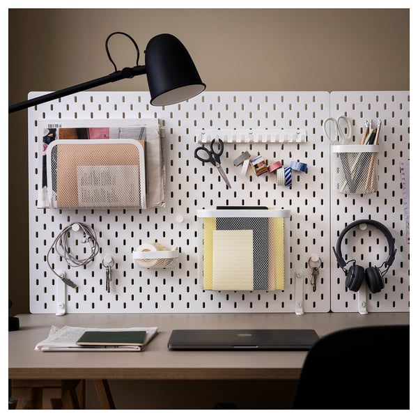 Scrivania con pannello forato bianco organizzato con lampada nera, cestini di metallo, forbici, cuffie, chiavi e altre forniture per ufficio.