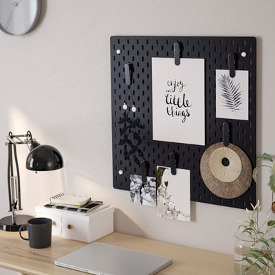 Scrivania con pannello forato SKÅDIS con foto e appunti; laptop, lampada, tazza e pianta sulla scrivania.