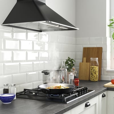 Cucina moderna con piano cottura a gas nero SKÅLAN, piastrelle bianche, piante e utensili da cucina. Cappa nera sopra, pentole sul fornello, assi di legno e barattoli.