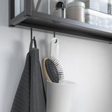 Il piccolo contenitore bianco SKATTÅN è appeso sotto il ripiano e contiene una spazzola per capelli. Asciugamano grigio e articoli da toeletta sullo scaffale.