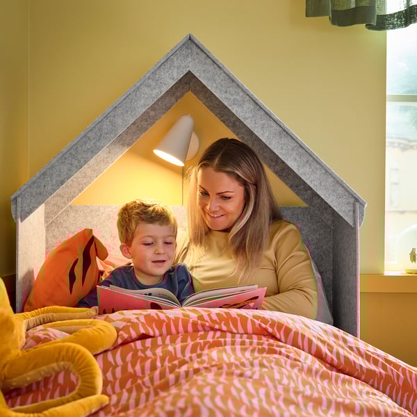 Una donna e un bambino leggono a letto, illuminati da una lampada montata su una testiera di legno, creando un'atmosfera accogliente.