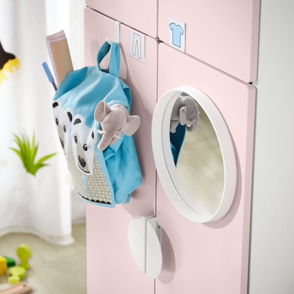 Armadio per bambini rosa chiaro con zaino blu e peluche sui ganci sinistri, specchio rotondo sulla porta destra.