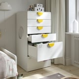 Cassettiera bianca per bambini con cassetti, maniglie gialle e piedini regolabili. I cassetti superiori mostrano la conservazione dei vestiti per neonati/bambini.