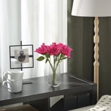 Scena accogliente della scrivania: dalie SMYCKA rosa brillante in vaso trasparente, tazze bianche, lampada moderna, opera d'arte minimalista. I fiori si stagliano alti tra decorazioni semplici.