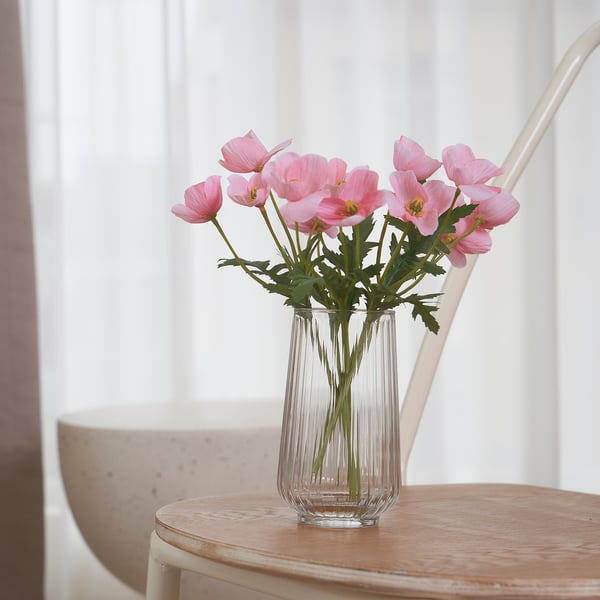 Un vaso chiaro a coste con fiori artificiali rosa posto su un tavolo di legno vicino a una finestra. I fiori portano colori vivaci e decorazioni allo spazio.