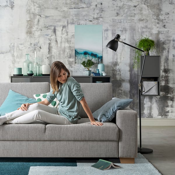 La donna si rilassa sul divano grigio SÖRVLEN, design minimalista, tessuto strutturato, cuscino decorativo, soggiorno moderno con arte astratta.