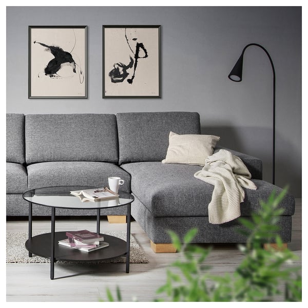 Soggiorno moderno con divano grigio SÖRVLEN, tavolino in vetro, stampe artistiche astratte, lampada da terra, pianta verde, comodi cuscini e coperta.