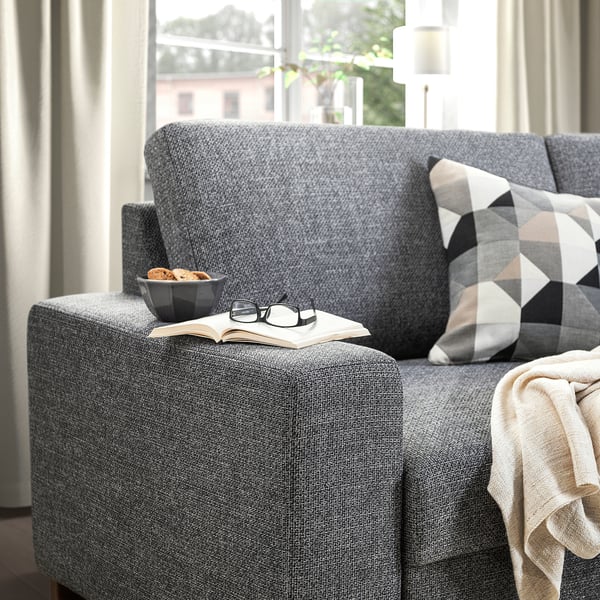 Divano grigio SÖRVALLEN vicino alla finestra con cuscino, coperta, libro, bicchieri e snack.