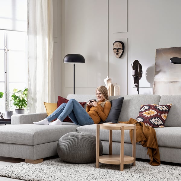 Donna rilassante su moderno divano grigio SÖRVLEN, accogliente soggiorno con luce naturale, cuscini colorati, tavolino in legno e lampada da terra.