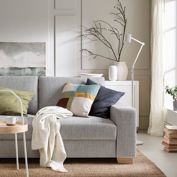 Moderno divano grigio a 3 posti con cuscini colorati, tavolino bianco, lampada e piante in una stanza luminosa.