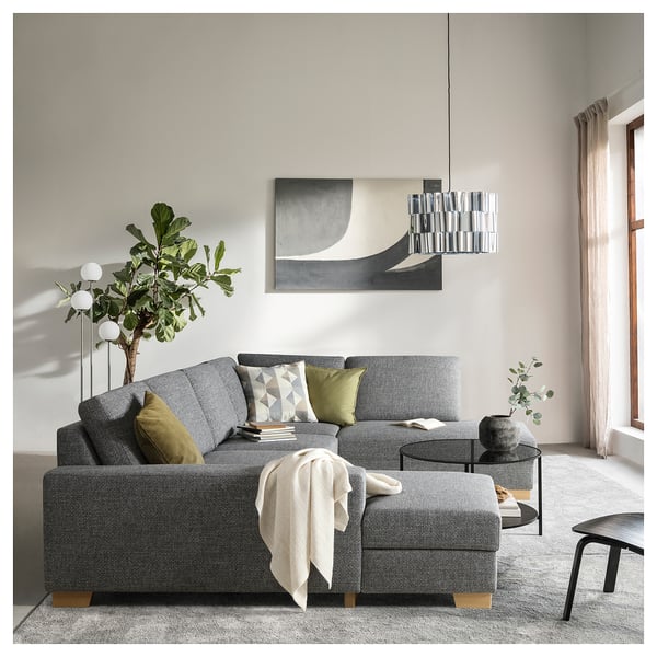 Soggiorno moderno con divano SÖRVALLEN grigio, cuscini verdi, elegante lampada a sospensione, arte murale astratta, coperta accogliente e piante in vaso.