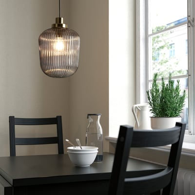 Angolo cucina moderno con grande lampada SOLKLINT a sospensione in ottone e vetro. Sedie nere, ciotola bianca, caraffa e pianta in vaso su un tavolo scuro vicino alla finestra.