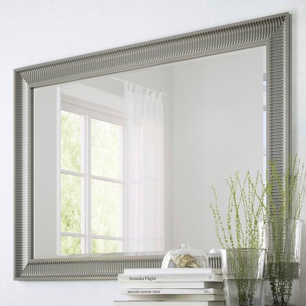 Grande specchio con opzioni di sospensione verticale e orizzontale, superficie riflettente, con pellicola di sicurezza. Mostrato sulla finestra riflettente a parete, accanto a piante e libri.