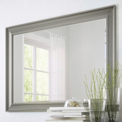 Grande specchio con opzioni di sospensione verticale e orizzontale, superficie riflettente, con pellicola di sicurezza. Mostrato sulla finestra riflettente a parete, accanto a piante e libri.