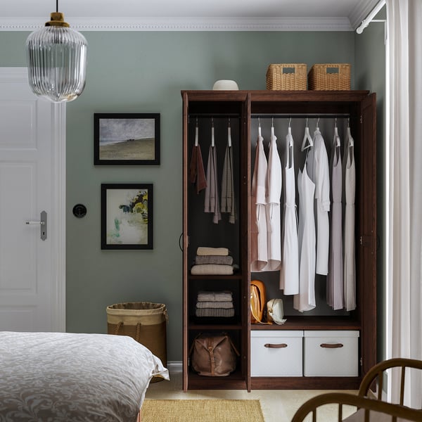 Armadio in legno con specchio, che espone vestiti e oggetti piegati. Due cestini in alto, borsa in basso. Montaggio a parete, parete verde, letto parziale.