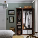 Armadio in legno con specchio, che espone vestiti e oggetti piegati. Due cestini in alto, borsa in basso. Montaggio a parete, parete verde, letto parziale.