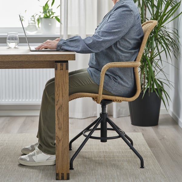 Persona che digita sul laptop in camicia grigia, pantaloni verdi, con i piedi sulla base della sedia di vimini.