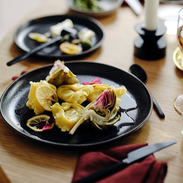 Piatti piani neri opachi con fette di carciofo e limone, distanziati su un tavolo di legno con coltelli e bicchieri.