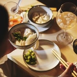 Due bicchieri da vino con bevande, ciotole di gnocchi e wasabi, bacchette su un piatto.
