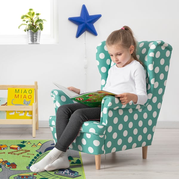 Ragazza che legge in sedia per bambini a pois verdi con libreria e pianta.